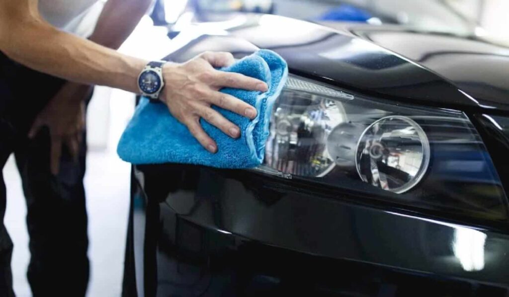 A person using a blue microfiber cloth to clean the headlight of a black car in a garage setting. The individual is wearing a watch and has their hand positioned on the cloth while focusing on the cleaning task.