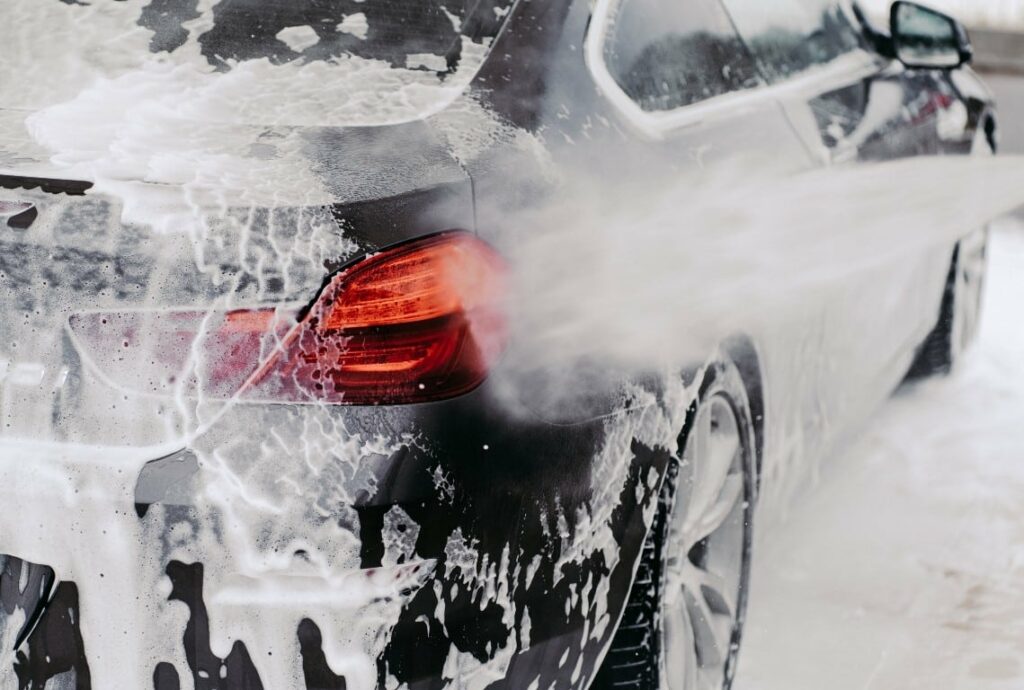 A black car being washed, covered in foam, with water spraying off its surface. The car's taillight is visible, highlighted by the foam surrounding it.