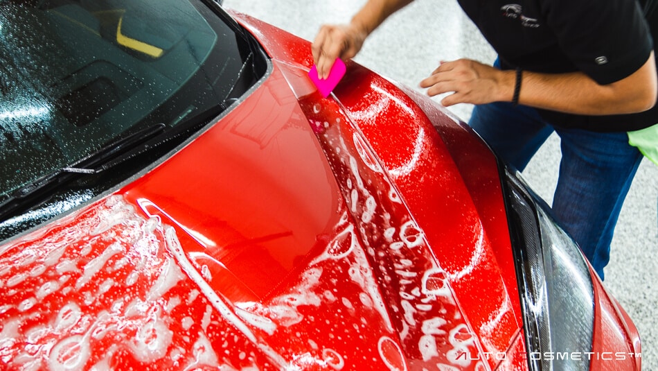 A person using a pink applicator to apply ceramic coating on the hood of a red car, covered in a layer of soap suds, in a well-lit detailing garage.