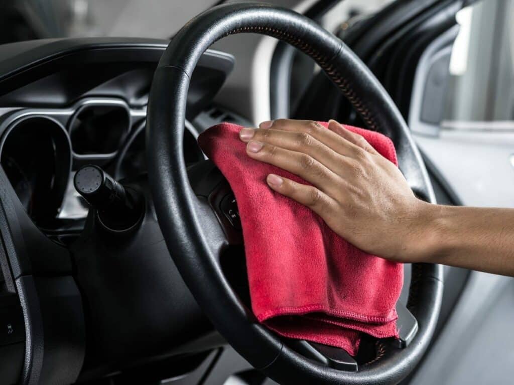 A person cleaning a steering wheel of a car with a red microfiber cloth. The interior of the vehicle is visible, showing the dashboard and controls.