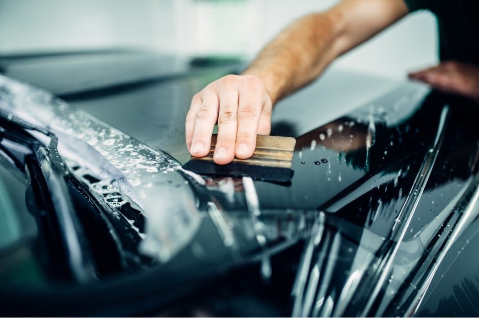 A close-up of a person using a squeegee to apply a protective film on the windshield of a black car. Water and soap residue are visible around the area being worked on.