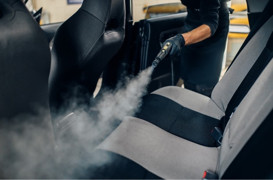 A person using a steam cleaner to sanitize the fabric seats of a car, with steam visibly emerging from the upholstery. The focus is on the back seat area, showcasing professional cleaning efforts.