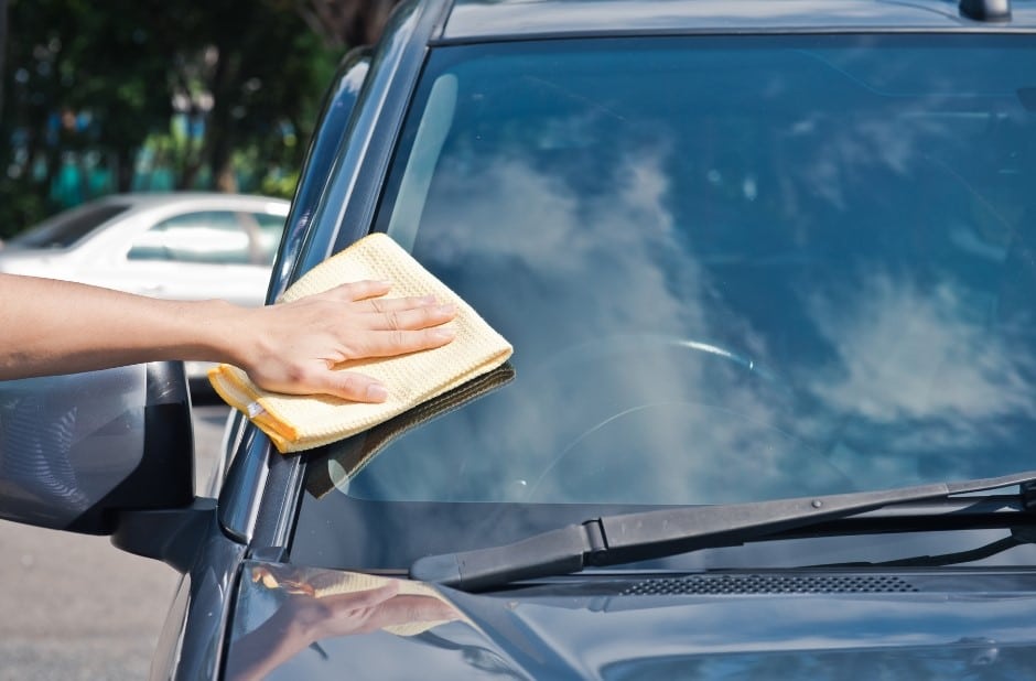 A person cleaning the windshield of a black car with a yellow microfiber cloth on a sunny day. The background shows other vehicles and blue sky.