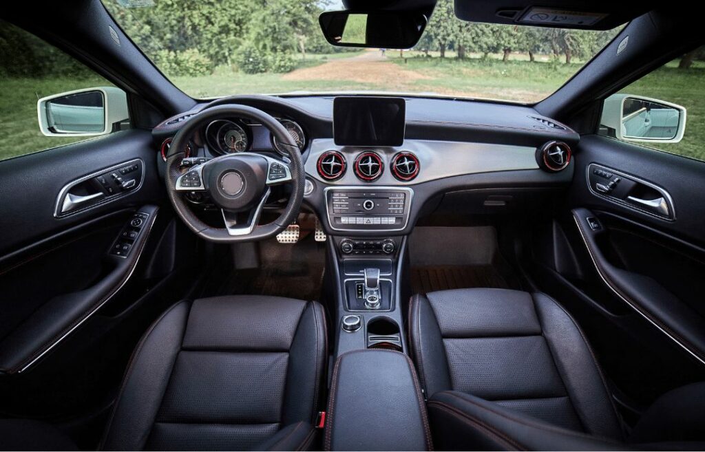 The interior of a luxury car featuring black leather seats, a modern dashboard with circular air vents, a digital display screen, and various controls. The view shows a grassy landscape through the windshield.