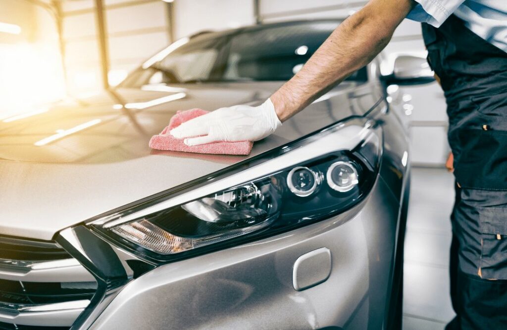 A person wearing a blue shirt and white gloves uses a red microfiber cloth to wipe the hood of a silver car in a well-lit garage.