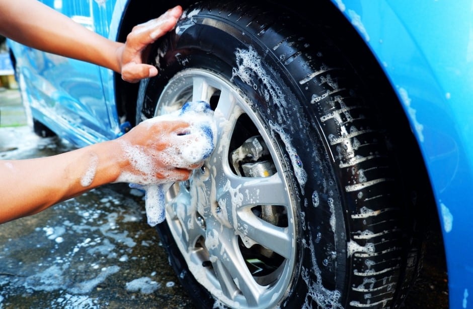 A person cleaning a car tire with a soapy sponge, with bubbles visible on the tire and surface. The car is blue, and water is splashing around on the ground.