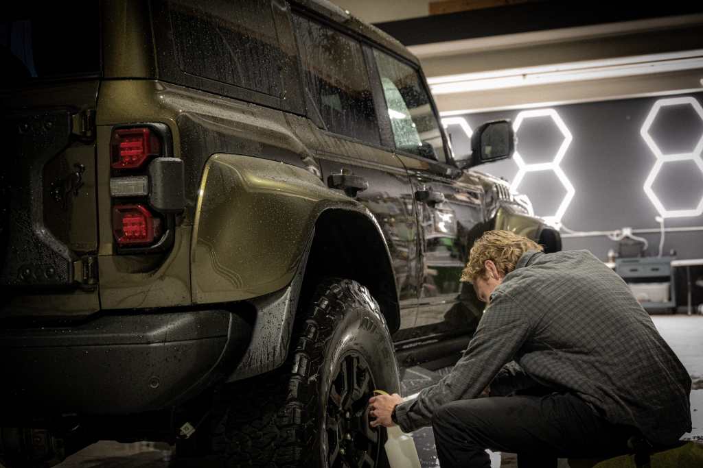 A person in a checkered shirt cleaning the wheel of a dark green SUV in an indoor setting with modern lighting.