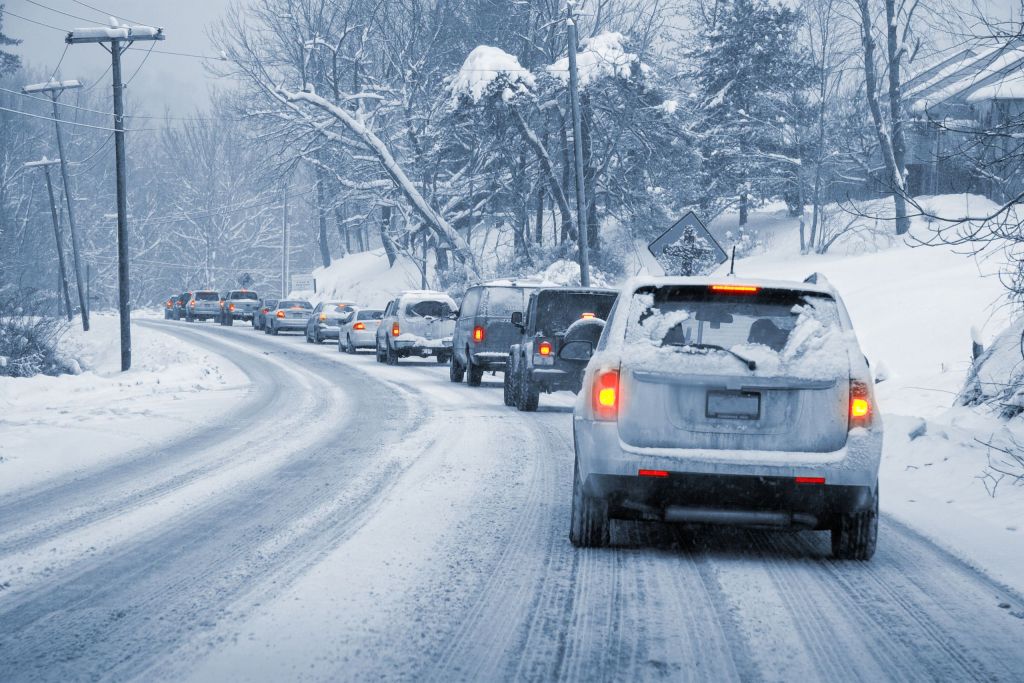 A line of vehicles navigating a snowy, winding road surrounded by trees. Snow covers the ground and the cars, with tire tracks visible in the snow. The scene is gray and overcast, indicating winter conditions.