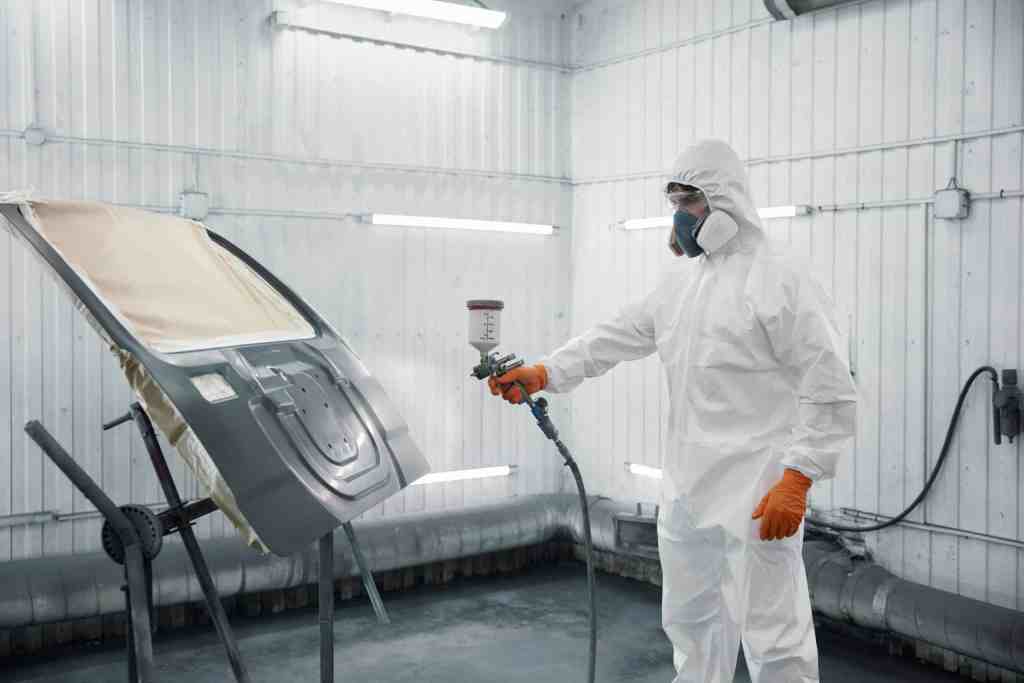 A person in protective gear spraying paint on a car hood in a well-lit workshop. The individual is wearing a full white suit, gloves, and a mask while using a spray gun. The background features white walls and industrial lighting.