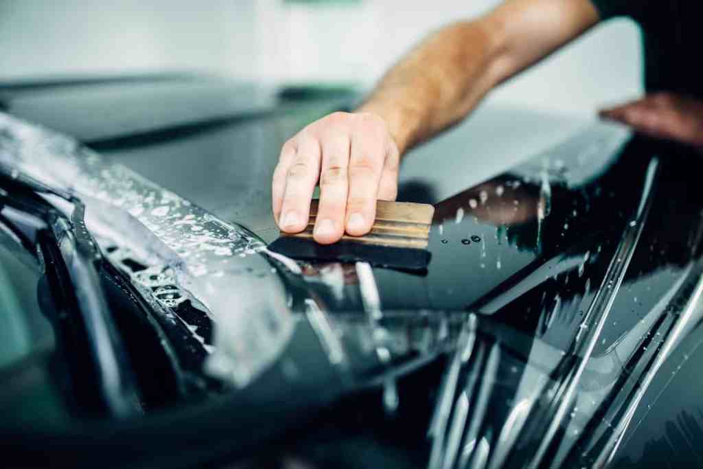 A close-up of a person's hand using a squeegee to remove water from a surface of a black car, with droplets and soap visible. The background is slightly blurred, indicating a car detailing or wash environment.