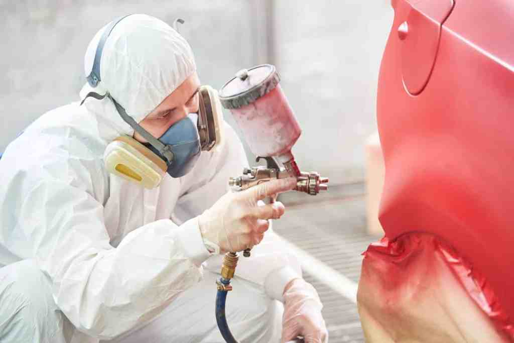 A person in a protective suit and mask using a spray gun to paint a red car in a workshop setting.
