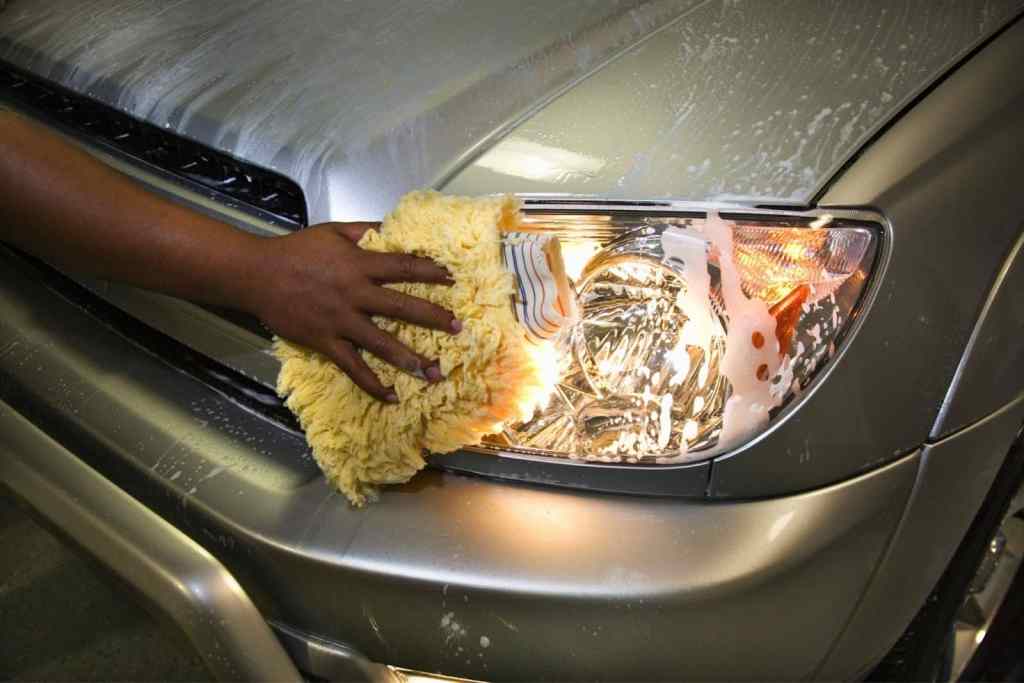 A hand using a yellow microfiber cloth to clean the headlight of a silver car, which is covered in soap and water.