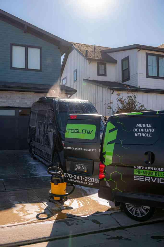 A black van labeled 'Autoglow' is being washed in a driveway in front of a modern two-story house. A pressure washer is placed nearby, and a second detailing van is visible, displaying text related to mobile detailing services.