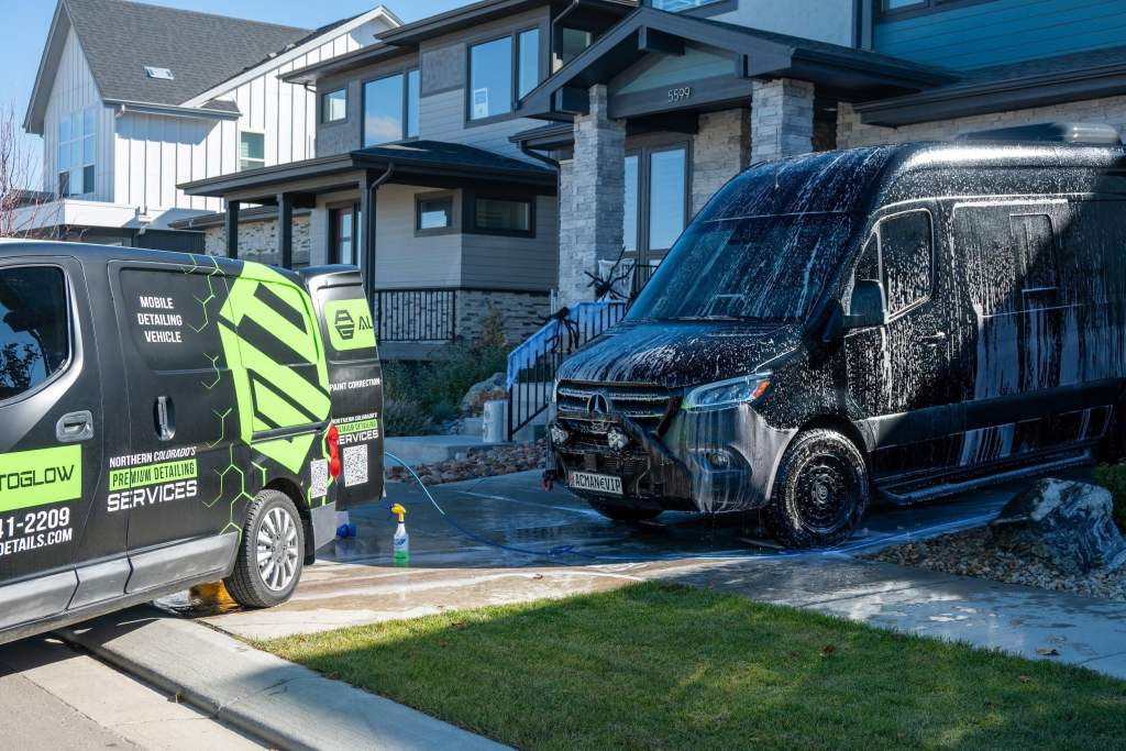 A black van covered in soap is parked in a driveway in front of a modern home. A detailing service vehicle with green graphics is nearby, showing its logo and contact information. The residential area features a clean lawn and stone steps.