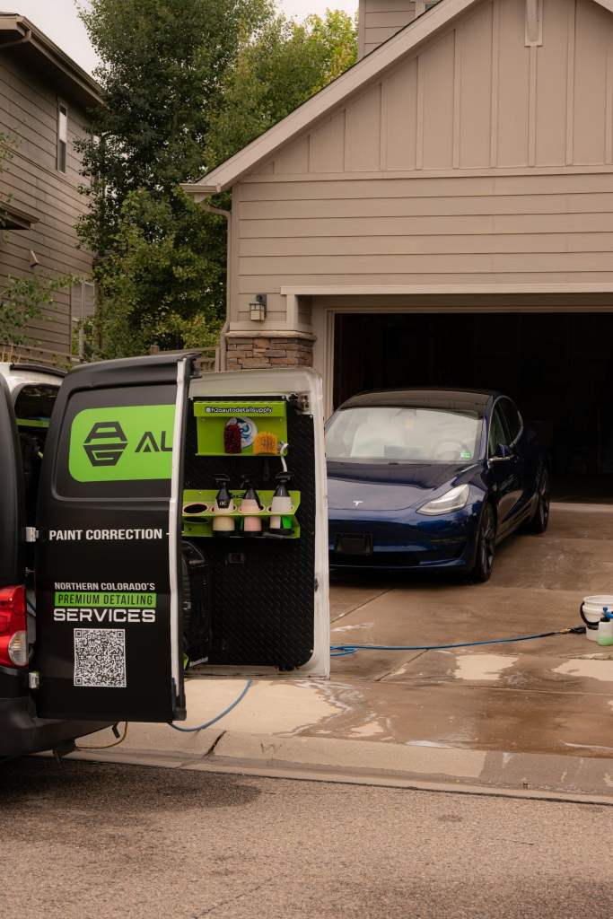 A parked dark blue Tesla electric vehicle is in a driveway in front of a light-colored garage. Open rear doors of a van display paint correction supplies and equipment marked with branding and contact information.