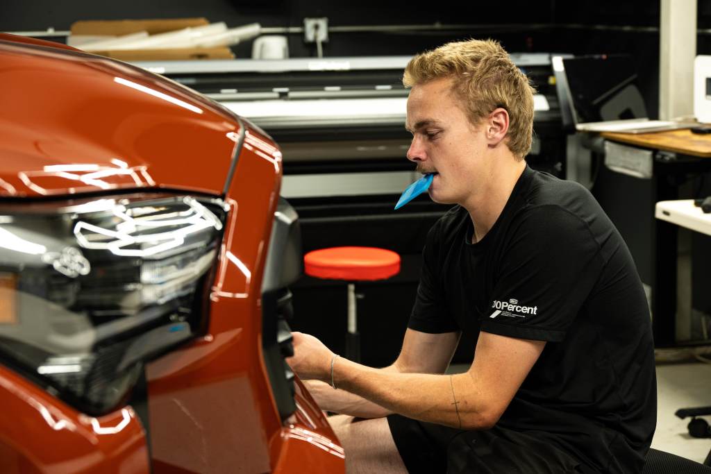 A young man with fair hair is seated next to an orange vehicle, focusing on a part under the headlight. He is holding a blue tool in his mouth while working with his hands. The background includes equipment and a chair, indicating a workshop setting.