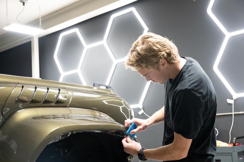 A young man applying a blue tool to the side of an olive green vehicle in a garage. He is focused on the detail work, and the background features a wall with hexagonal light patterns.