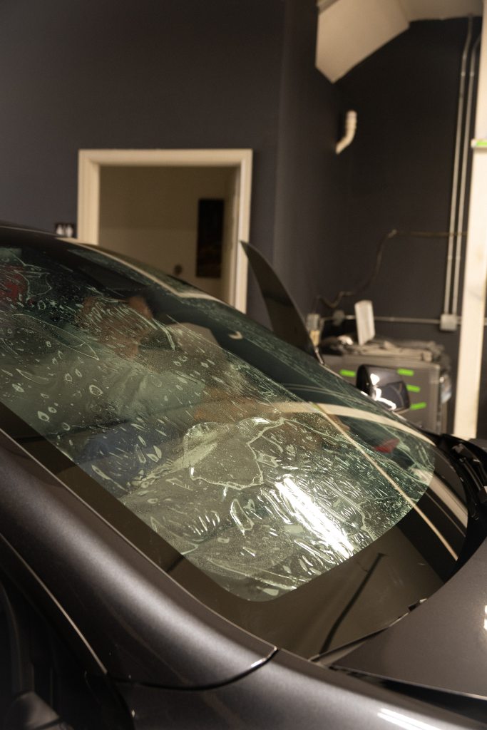 A close-up view of a car's windshield with protective film applied, showing bubbles and water droplets. The background includes a garage setting with a door frame visible and equipment in the distance.
