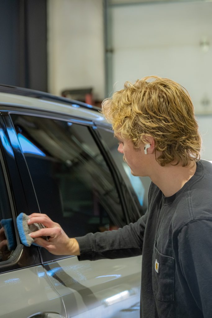 A young man with blond hair cleaning the window of a car using a cleaning tool. He is wearing a dark long-sleeve shirt and earbuds, focused on his task in a garage setting with blurred background elements.