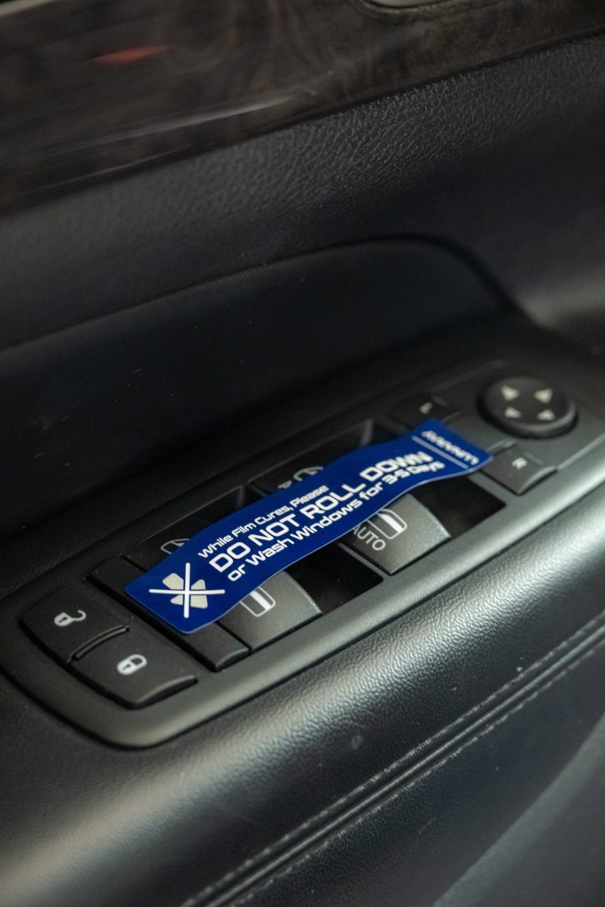 A close-up view of a vehicle's control panel featuring window controls and a blue warning sticker that reads, "DO NOT ROLL DOWN WINDOW." The panel is set against a black leather interior highlighting the vehicle's design.