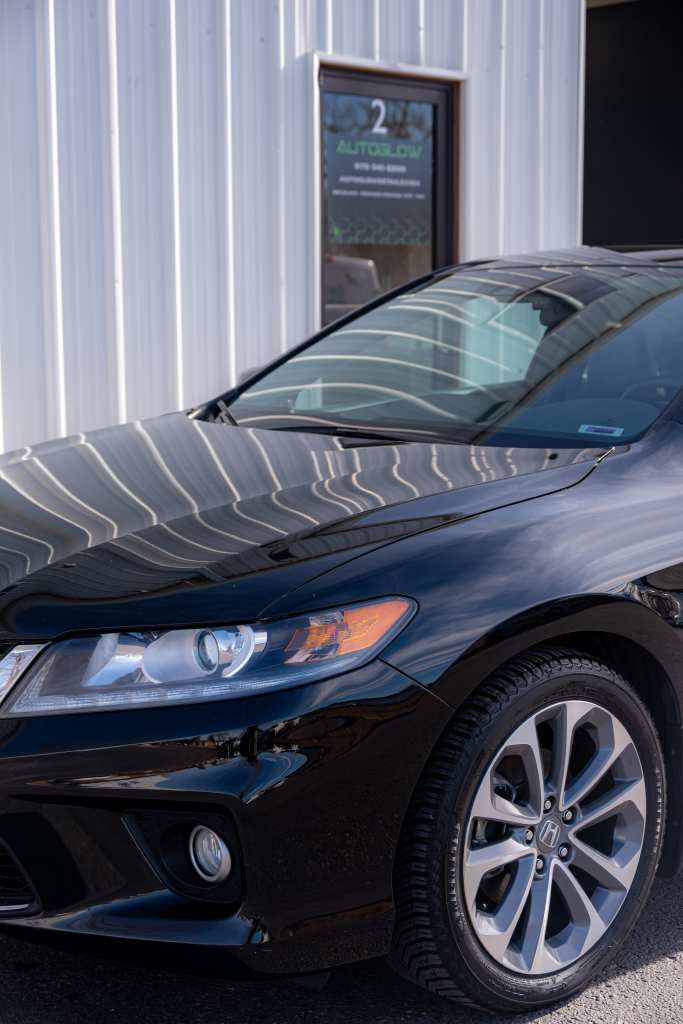 A black sedan parked near a building, showcasing its sleek profile and modern design. The vehicle's headlights and wheel are clearly visible, and reflections from the building's surface are noticeable on the car's hood.
