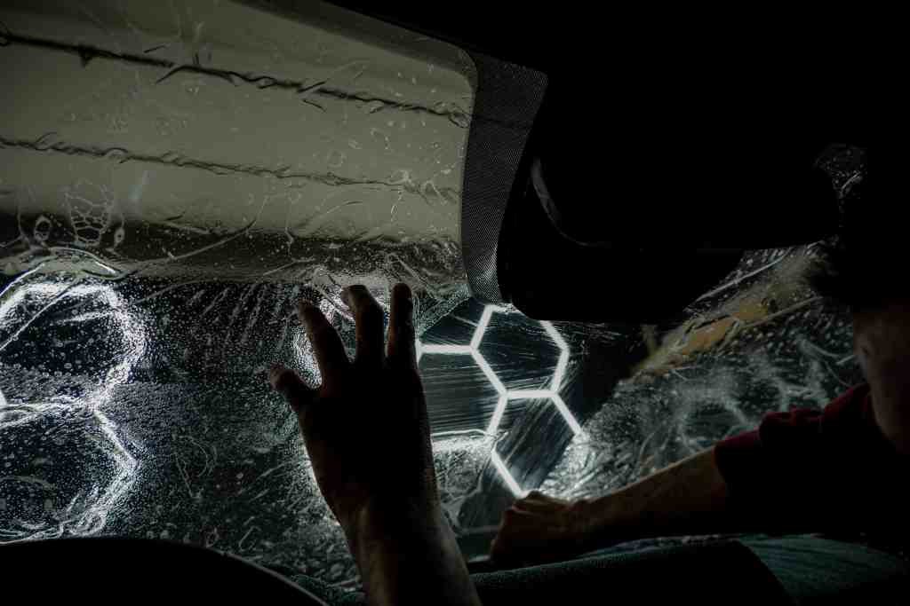 A close-up of a car window being cleaned with water and soap, creating bubbles and streaks. A hand is visible pressing against the glass from the inside, while a blurred figure is seen to the side, suggesting a car wash environment.