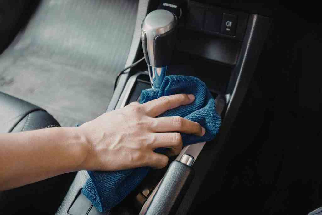 A hand using a blue microfiber cloth to clean the center console of a car. The console contains a gear shift and buttons, with a black leather interior visible in the background.