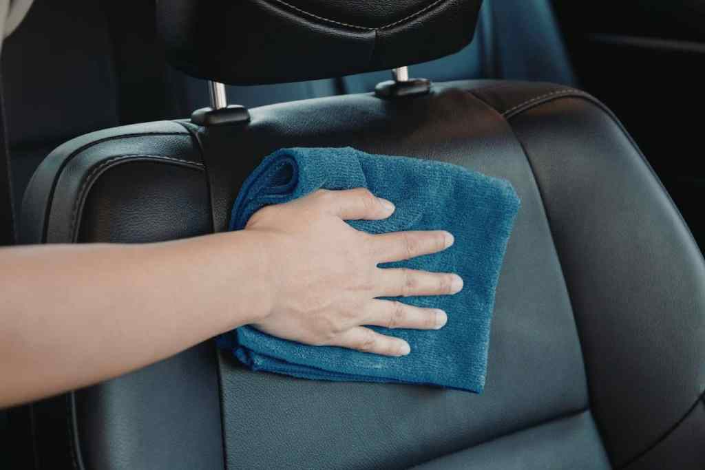 A hand holding a blue microfiber cloth, wiping the headrest of a black leather car seat. The interior of the car is visible, showcasing clean and polished surfaces.