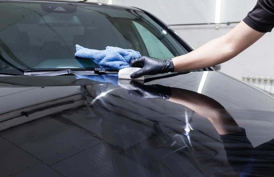 A person wearing a black glove using a cloth to polish the surface of a black car hood. Reflections of lights from the ceiling are visible on the glossy finish. The background shows a light-colored wall.