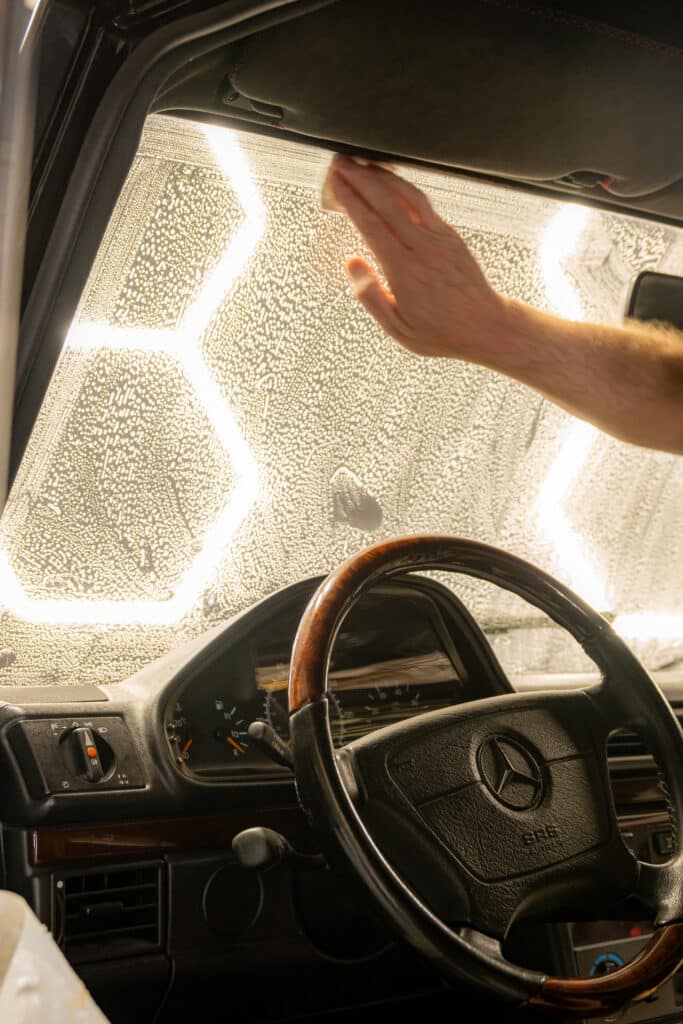 A person's hand wiping condensation from the windshield of a Mercedes-Benz car, with a wooden steering wheel and illuminated hexagonal patterns in the background.