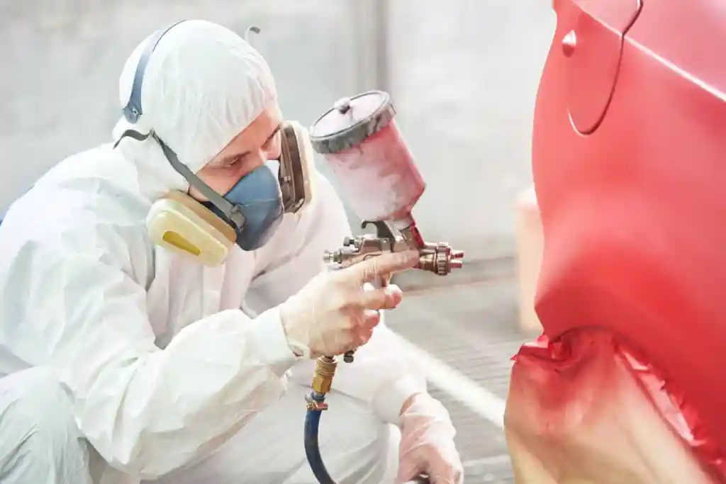 A person in a protective suit and mask using a spray gun to paint a red car in a workshop setting.