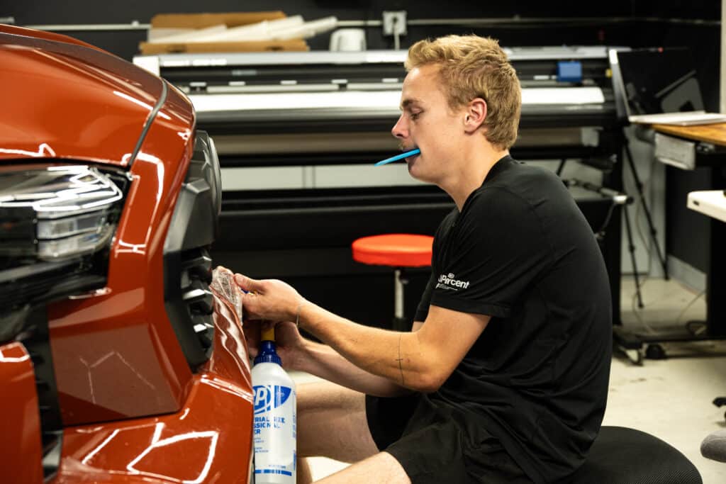 A young man sitting on a stool, cleaning the front grille of an orange car. He holds a cloth in one hand and a spray bottle in the other, with a toothpick in his mouth, in a workshop setting.