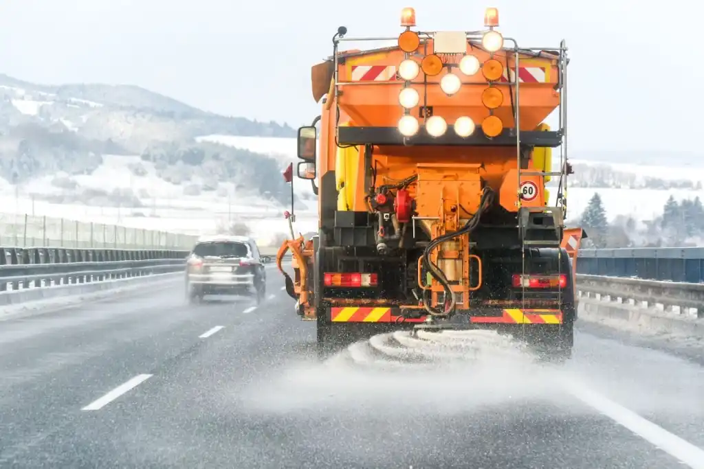 A snow plow truck spreading salt on a snow-covered highway, with a car driving in the opposite lane. Mountains are visible in the background under a cloudy sky.