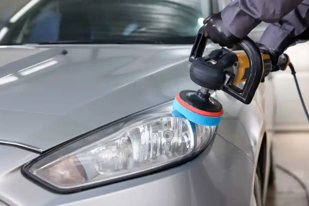 A person using a polisher on the headlight of a silver car in a garage setting. The polisher has a blue and red pad, and the environment is well-lit, showcasing the car's glossy surface.
