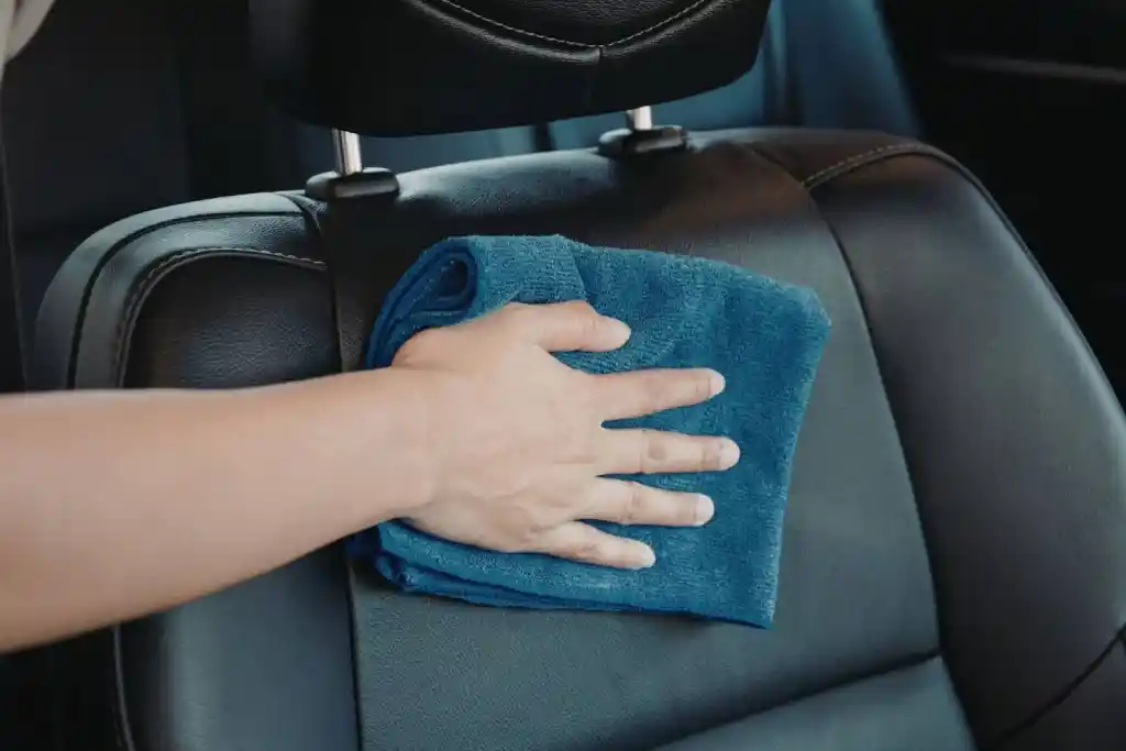 A hand holding a blue microfiber cloth, wiping the headrest of a black leather car seat. The interior of the car is visible, showcasing clean and polished surfaces.