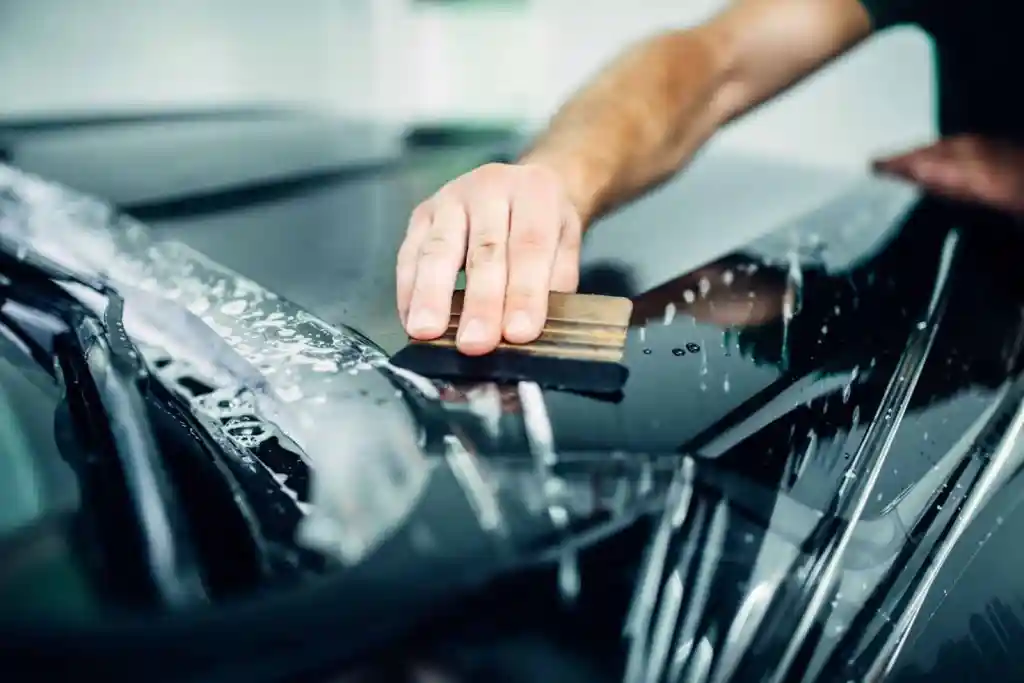 A close-up of a person's hand using a squeegee to remove water from a surface of a black car, with droplets and soap visible. The background is slightly blurred, indicating a car detailing or wash environment.