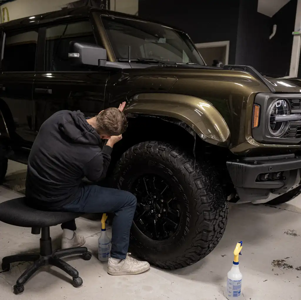 A person crouched on a swivel chair, inspecting the wheel arch of a dark green off-road vehicle in a garage. Cleaning supplies are placed on the floor nearby.